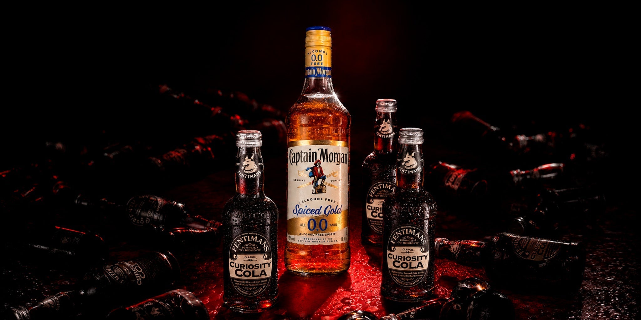 Captain Morgan Spiced Gold 0.0 alcohol-free spirit bottle with Fentimans Curiosity Cola bottles, styled in a dark dramatic bar scene with red lighting and party mood.