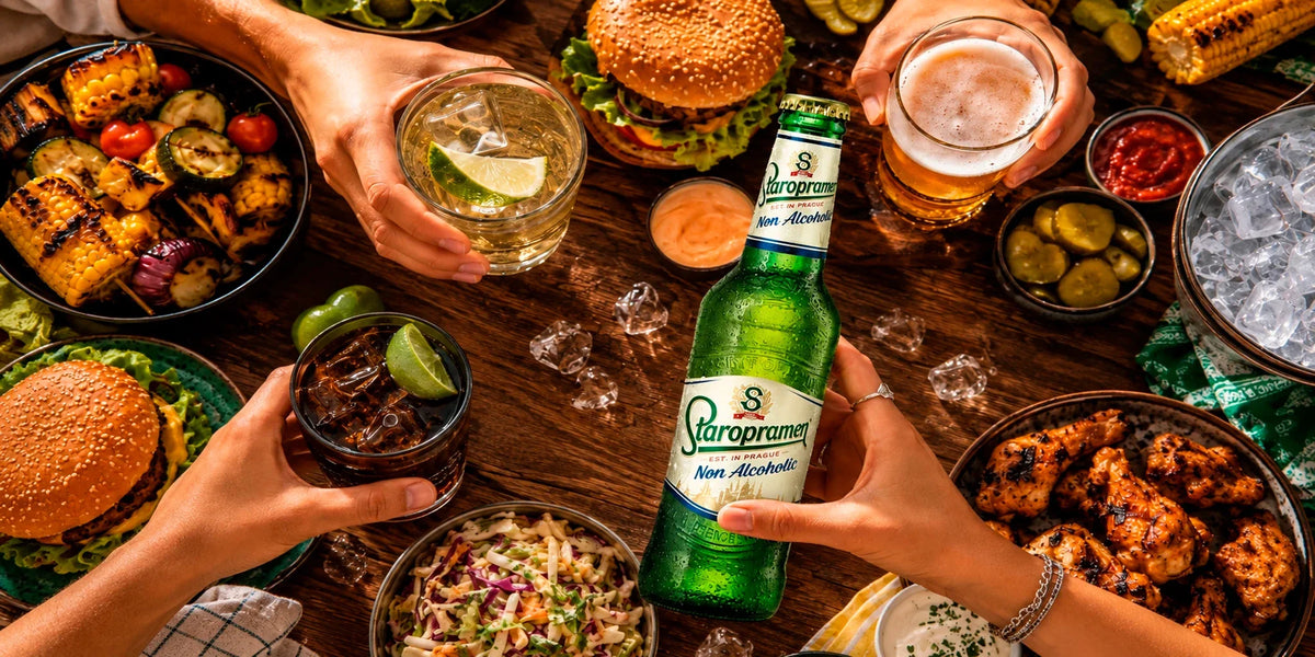 Staropramen Non-Alcoholic beer bottle shared at a lively BBQ table with burgers, grilled corn, chicken wings, pickles, ice and mixed drinks in a casual summer gathering.