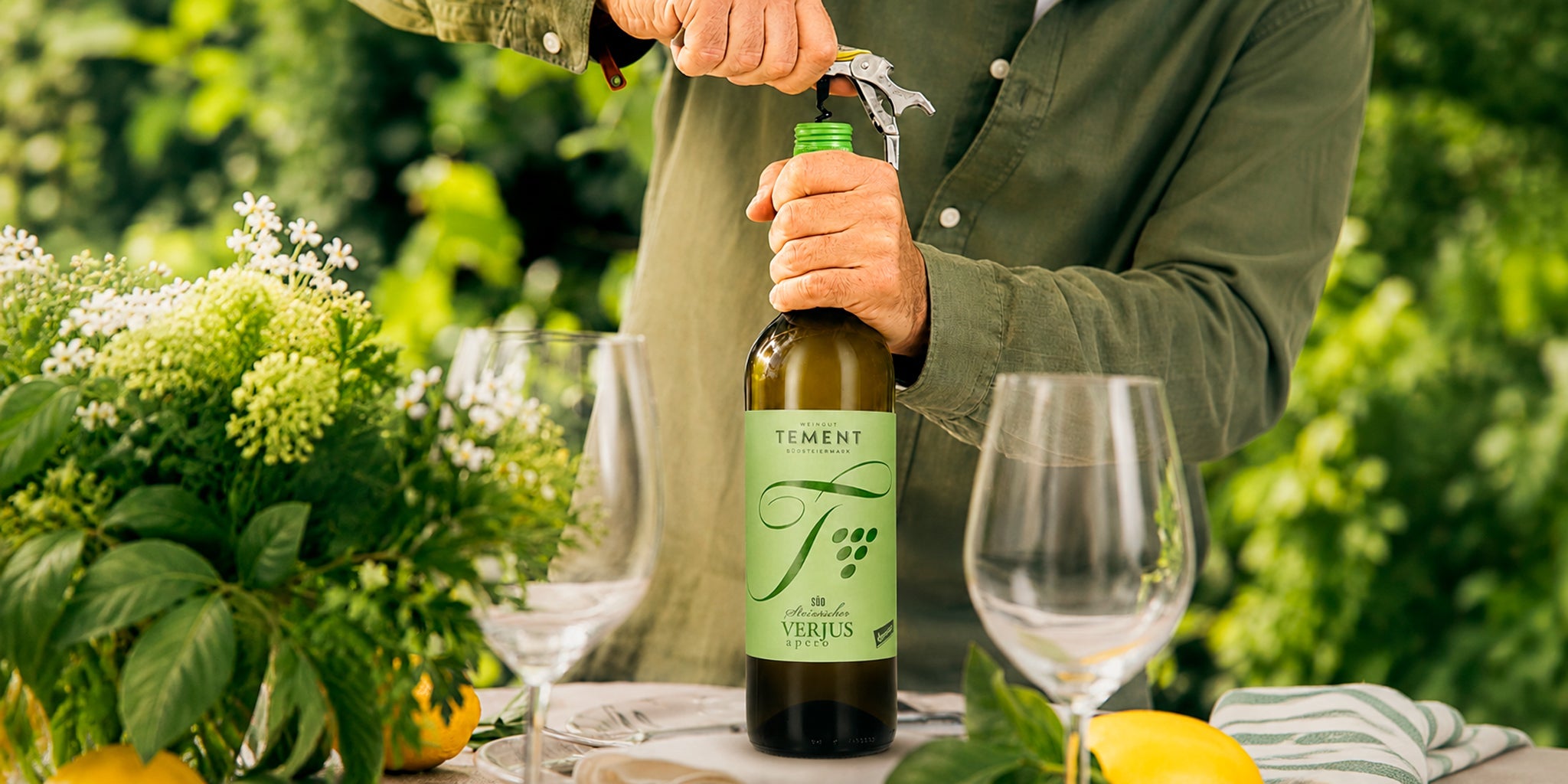 Tement Südsteirischer Verjus Apéro bottle being opened at a sunny garden table with wine glasses, fresh greenery and citrus, styled as a refined non-alcoholic aperitif for elegant outdoor serving.